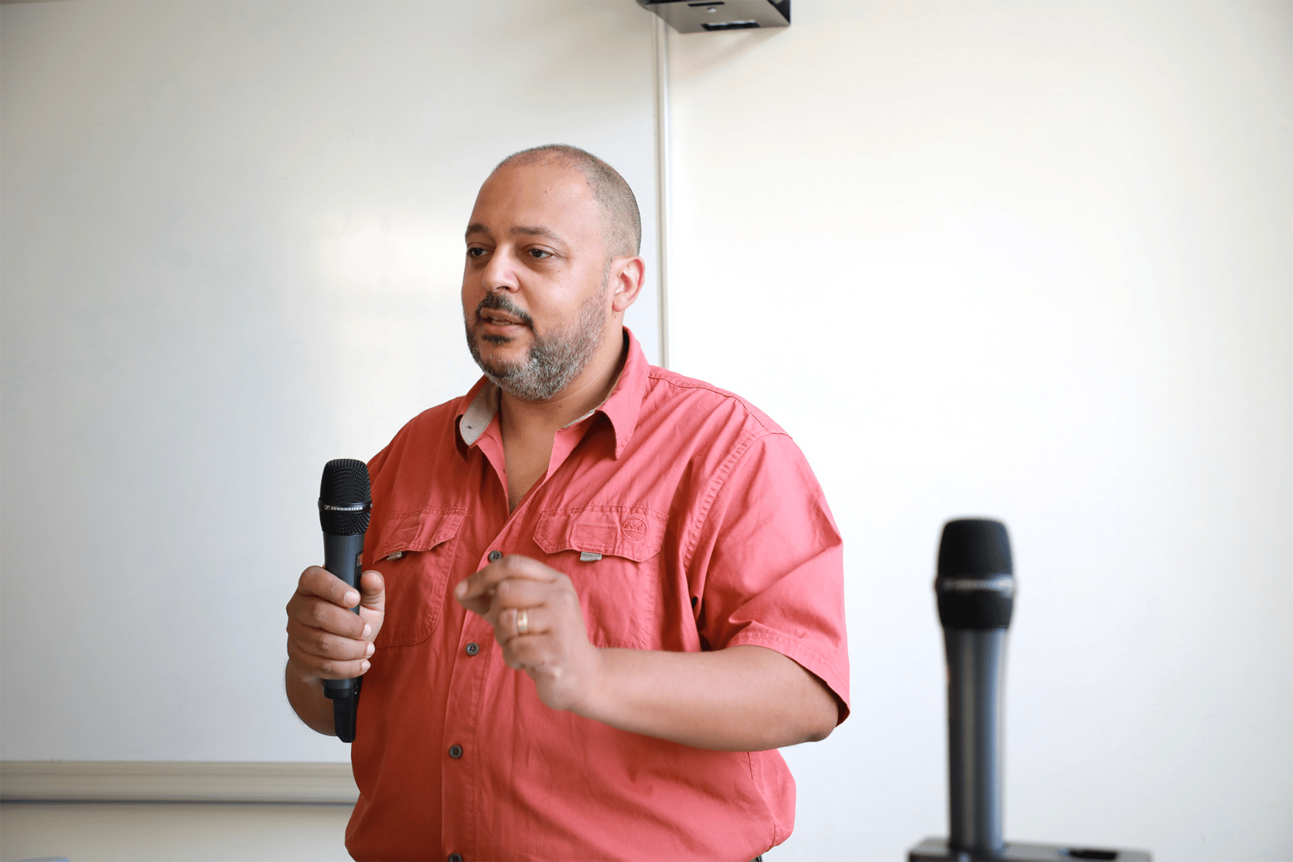 Man in a red shirt holding a microphone and speaking in a classroom setting.