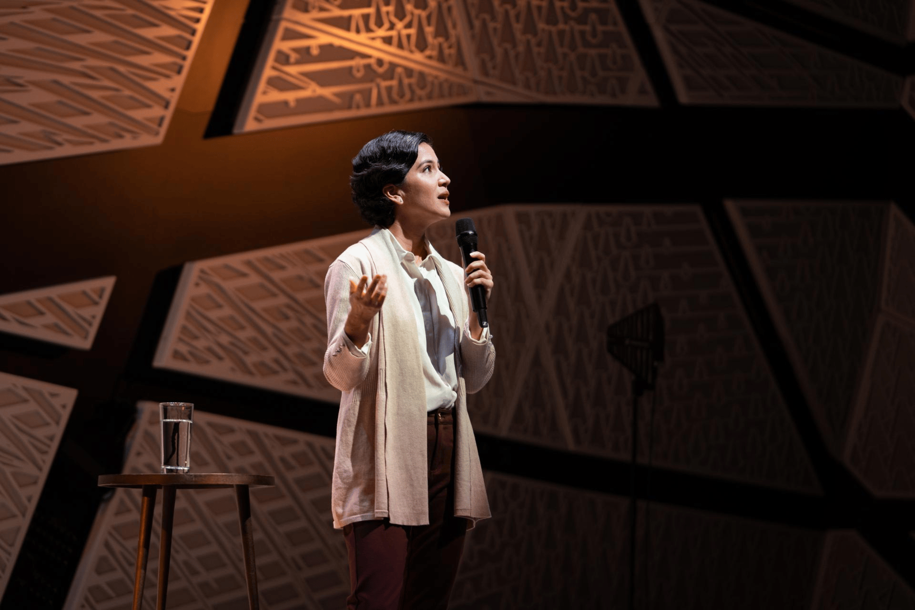 Person speaking into a microphone on stage with geometric background.