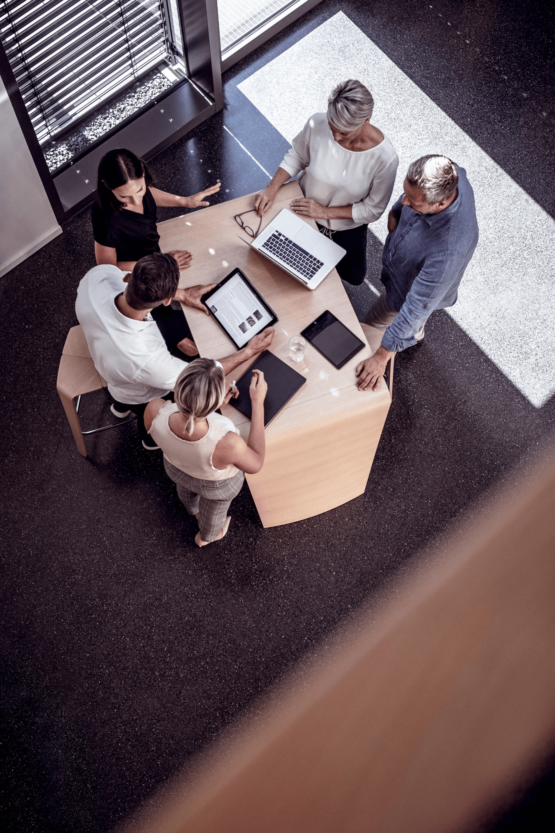 Group of people collaborating around a table with laptops and tablets in a modern office setting.