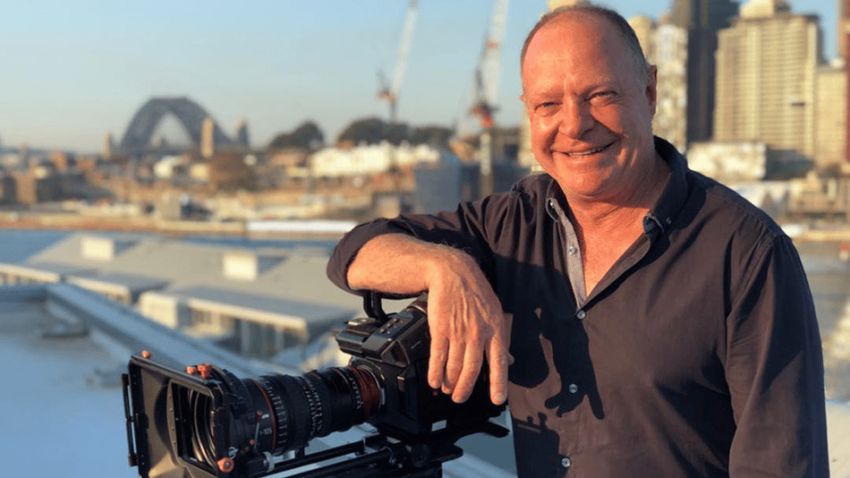 Man smiling with a professional camera on a rooftop with a cityscape and bridge in the background.