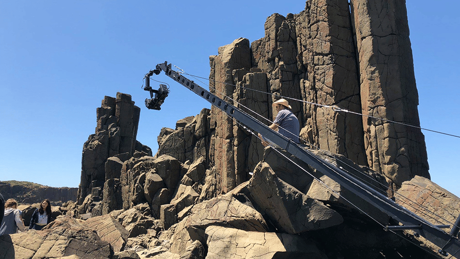 Film crew operating camera crane on rocky terrain with tall rock formations in the background.