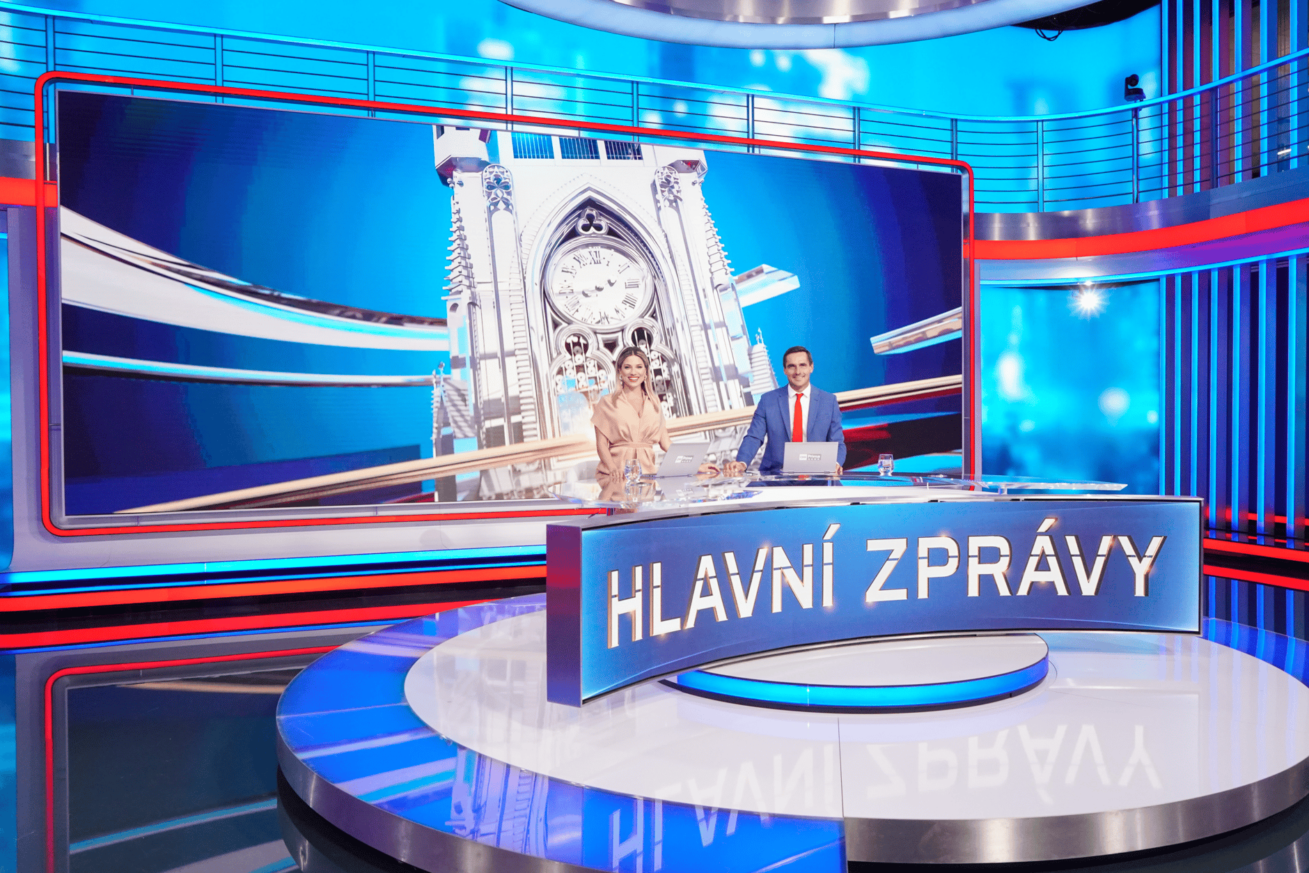 News anchors in a modern TV studio with a large digital screen displaying a clock tower.