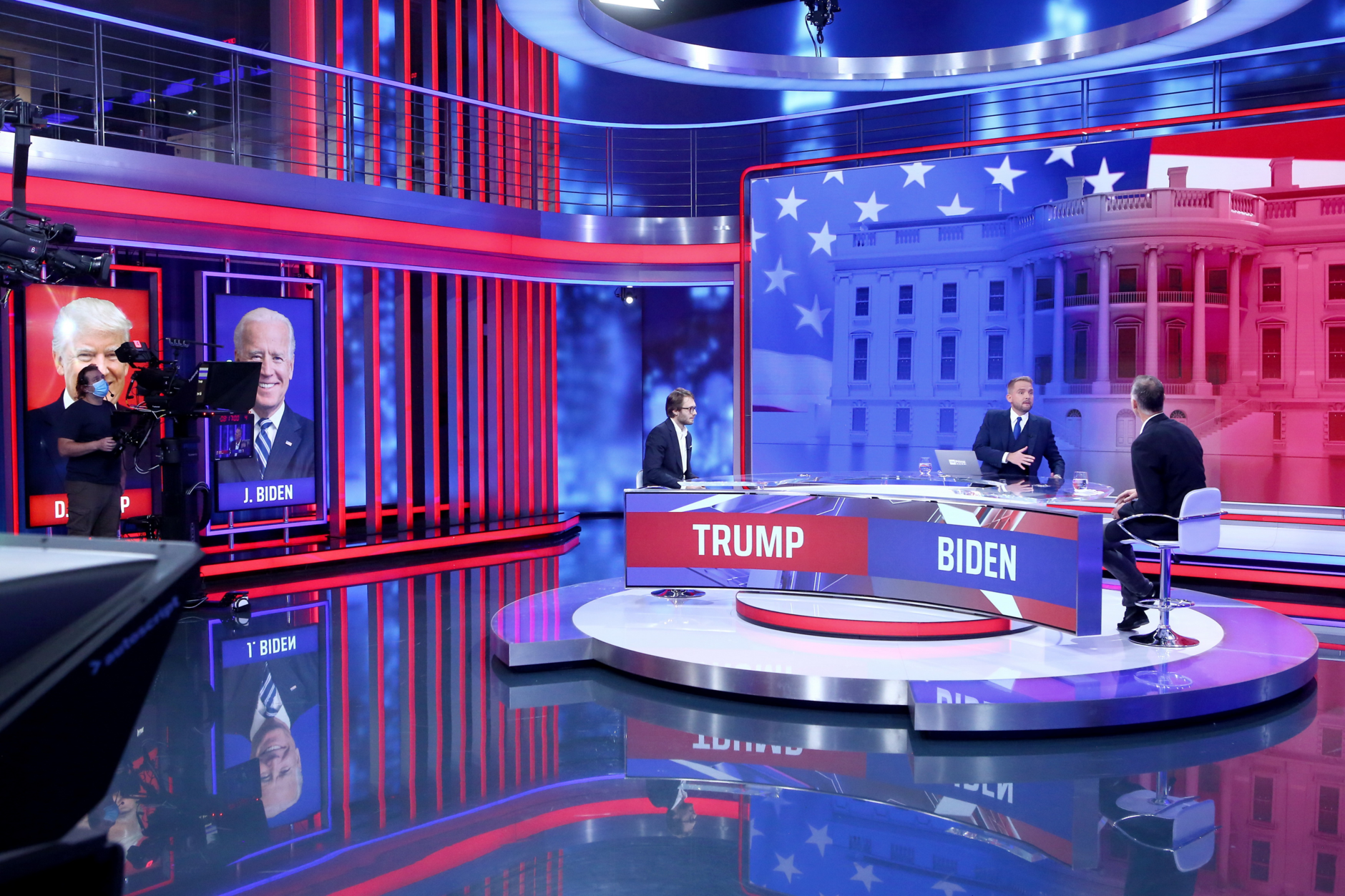 Television studio set with hosts discussing Trump and Biden in front of large screens displaying their images.