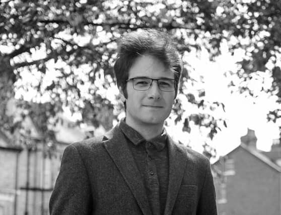 Man wearing glasses and a suit jacket standing outdoors with trees and buildings in the background.