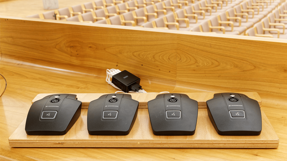 Four Sennheiser wireless conference microphones on a wooden desk in an empty auditorium.