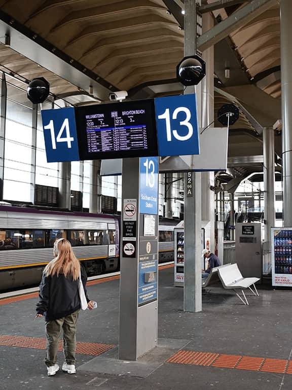 Train station platform with digital departure boards and a waiting passenger.
