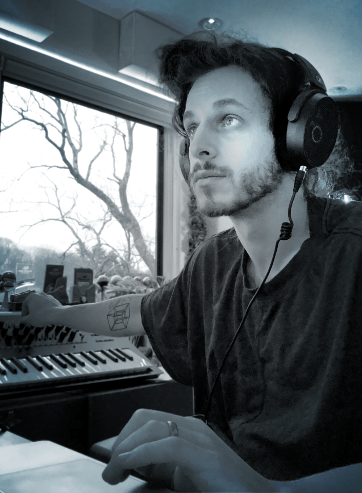 Man wearing headphones working on a music production setup with a keyboard in front of a window.