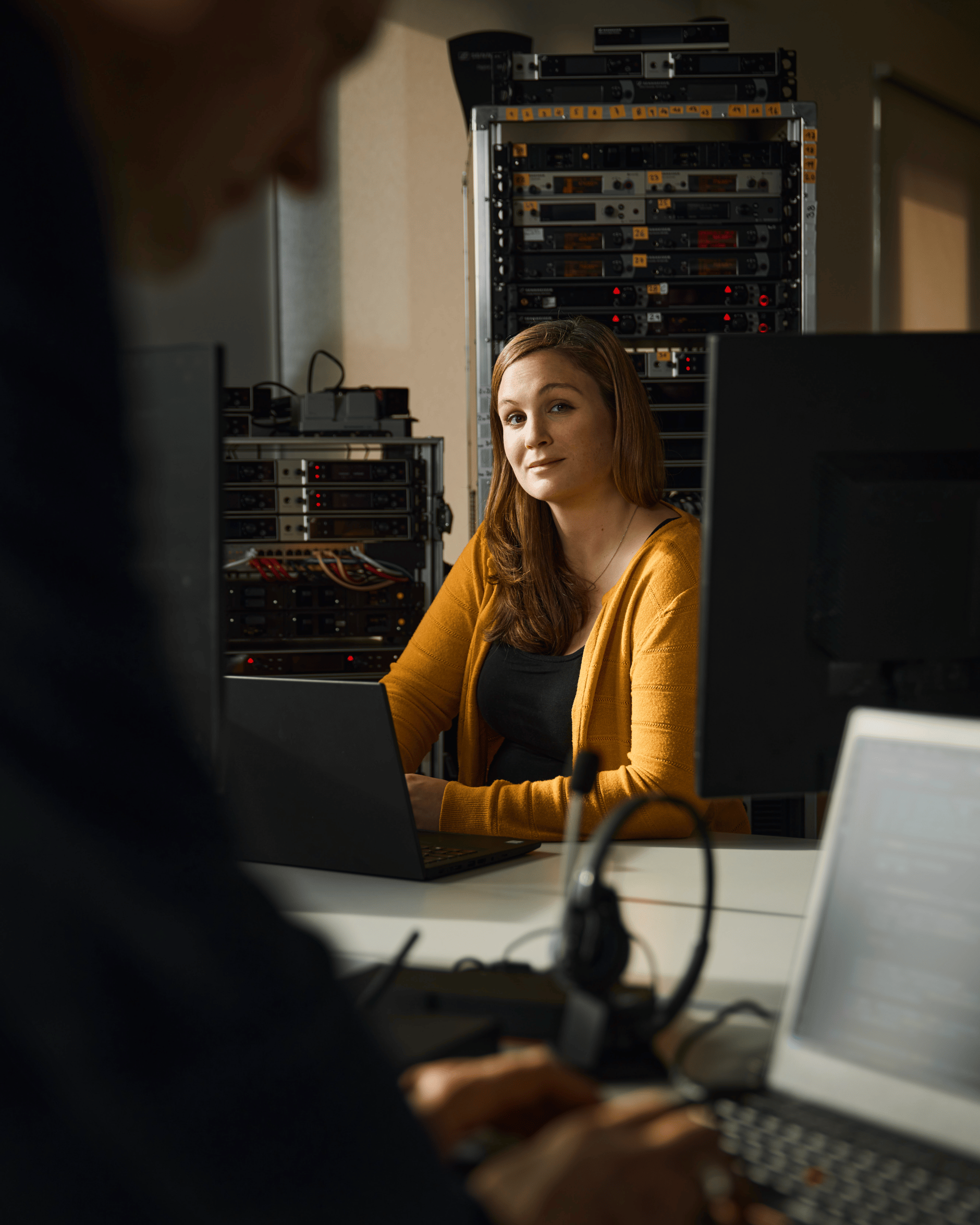Woman working on a laptop in a server room with network equipment in the background.
