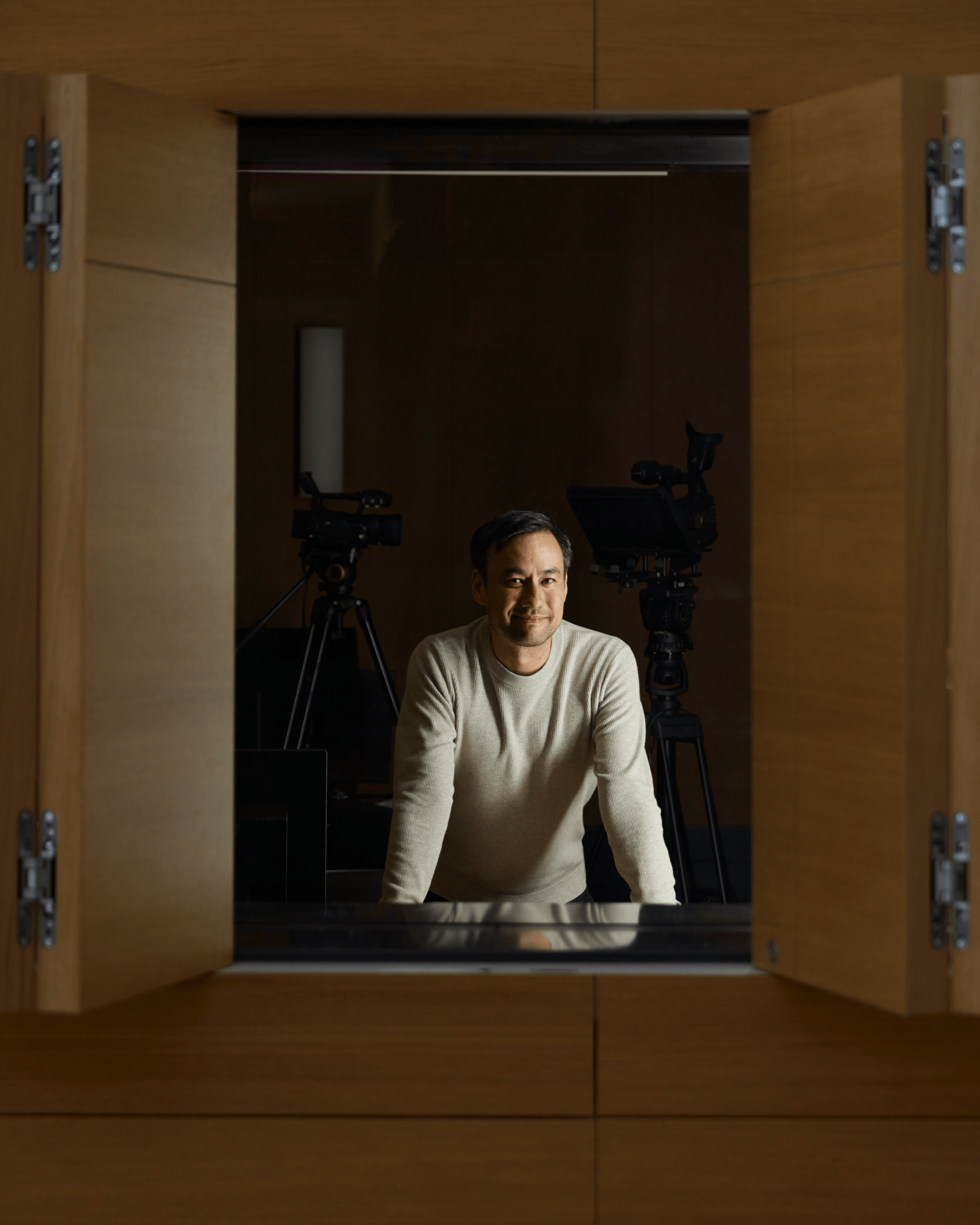 Man in a white sweater standing in front of professional video cameras in a wooden room.