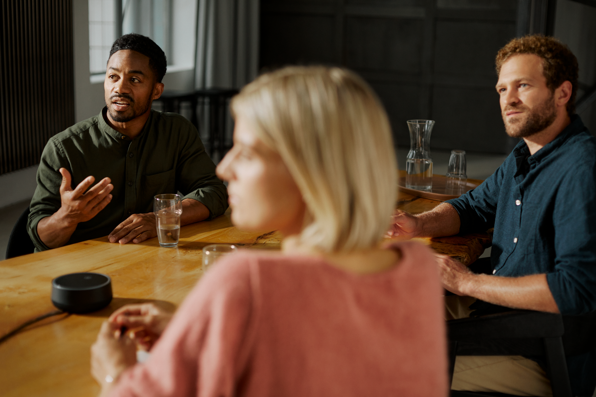 Two man and a woman having a meeting and sitting around a table with water glasses and a speaker on it 