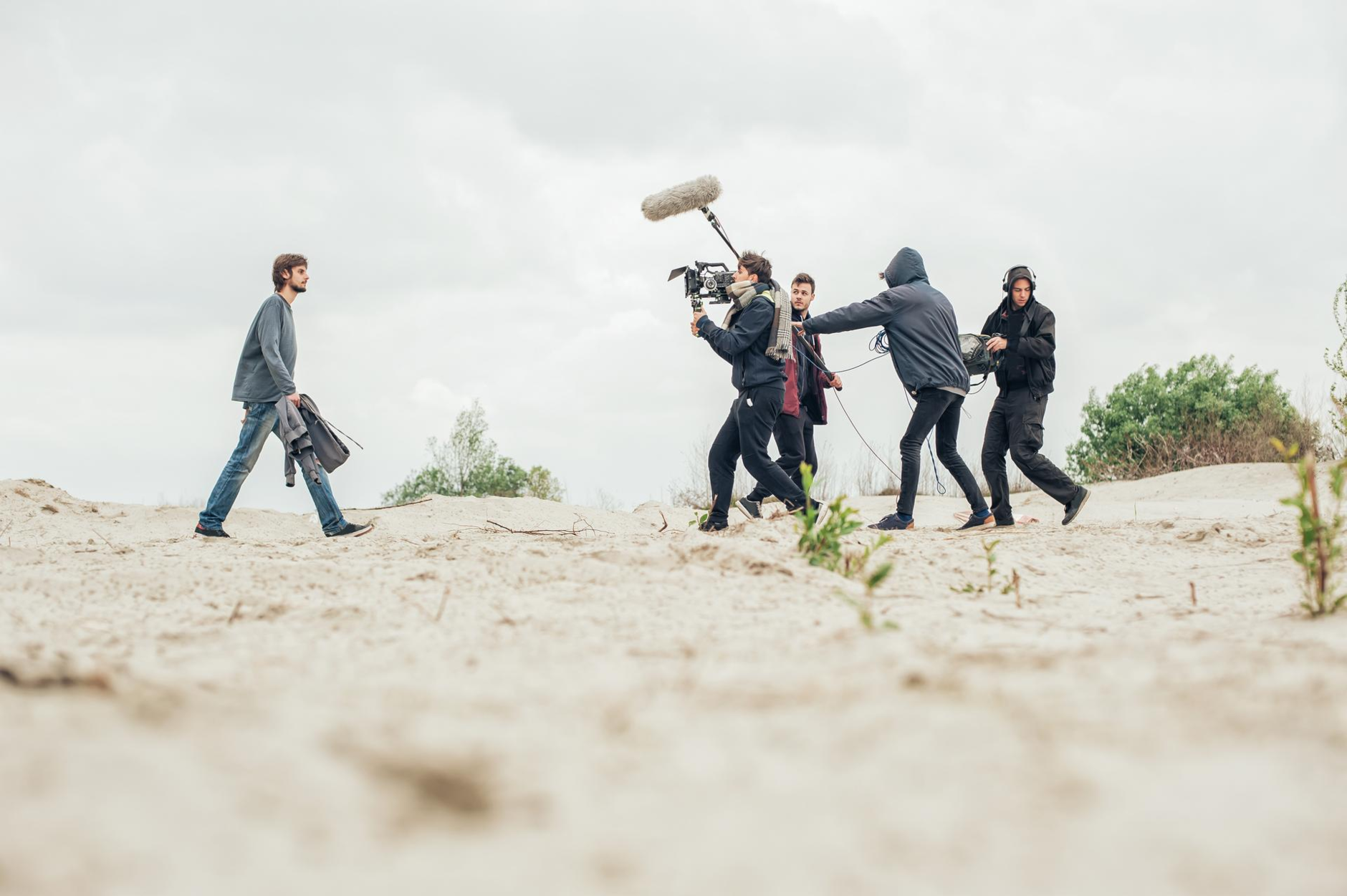 A small Film Crew of four men and an acteur are walking along a beach with their equipment, shooting a video or film 