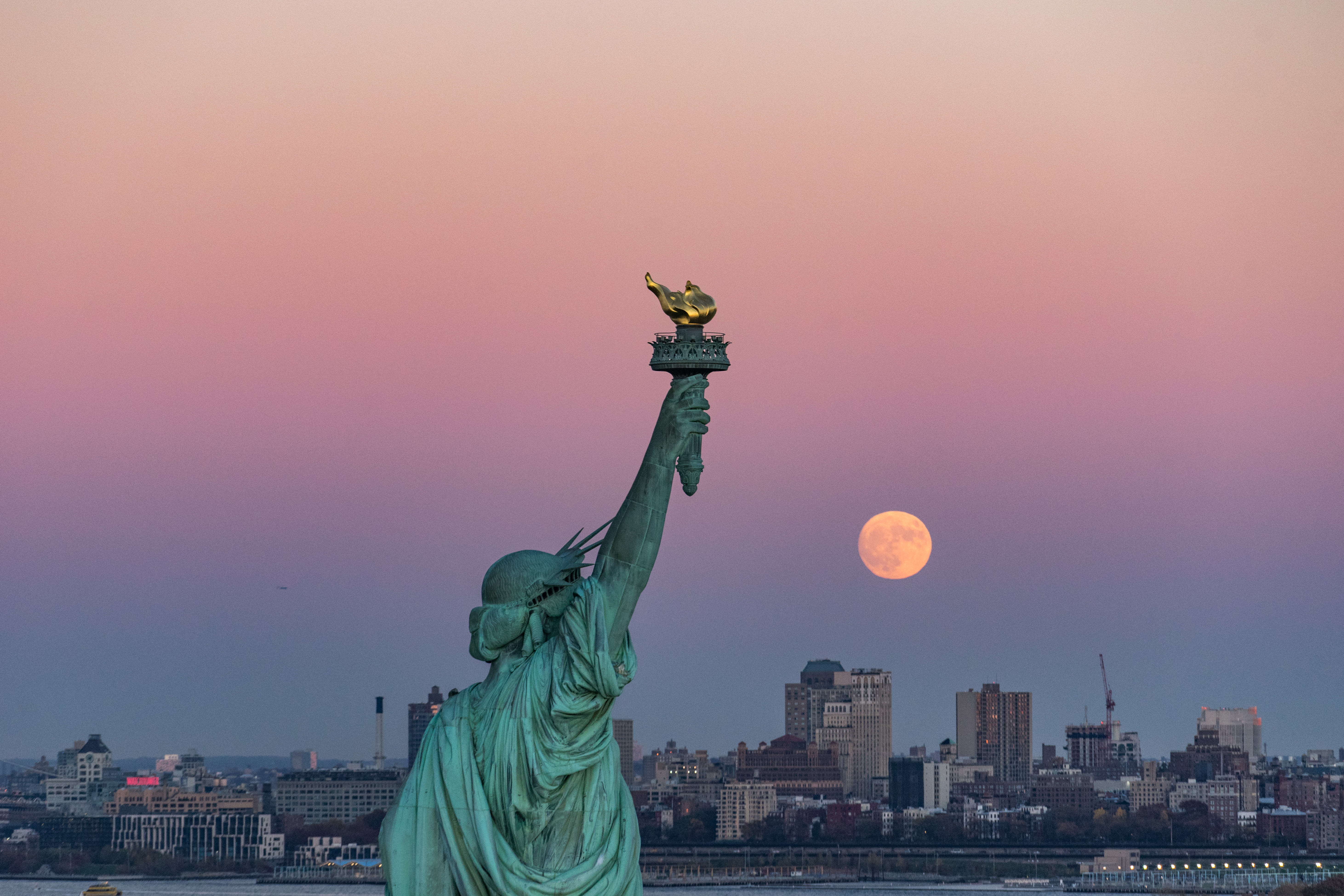 Statue of Liberty with New York City skyline at sunset and full moon.