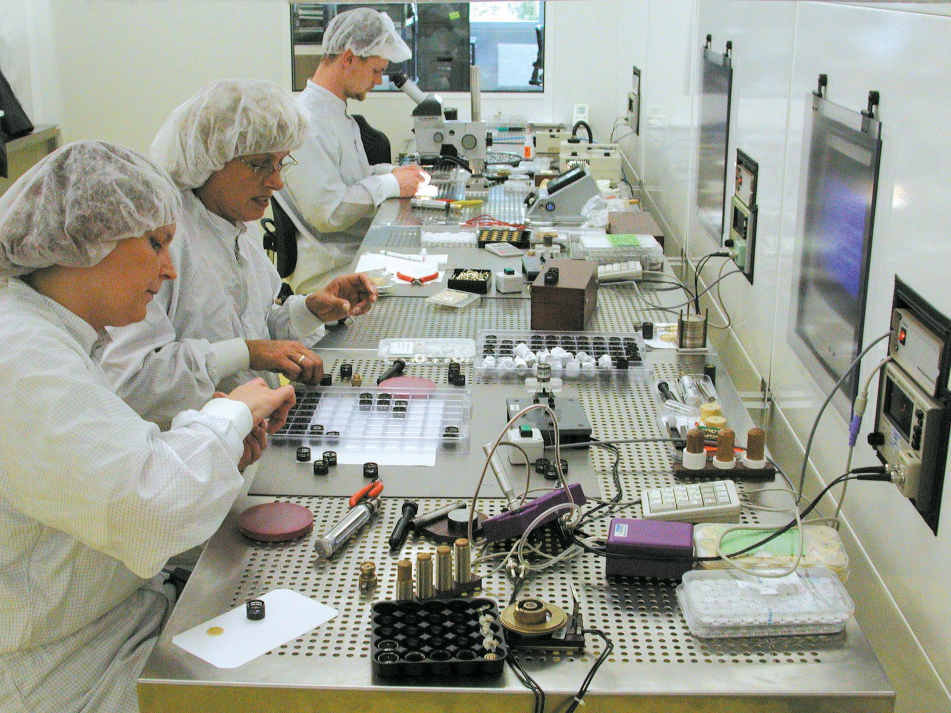 Two women and a man with smocks and hair protection sitting on a long workbench with several tools and a microscope on it 