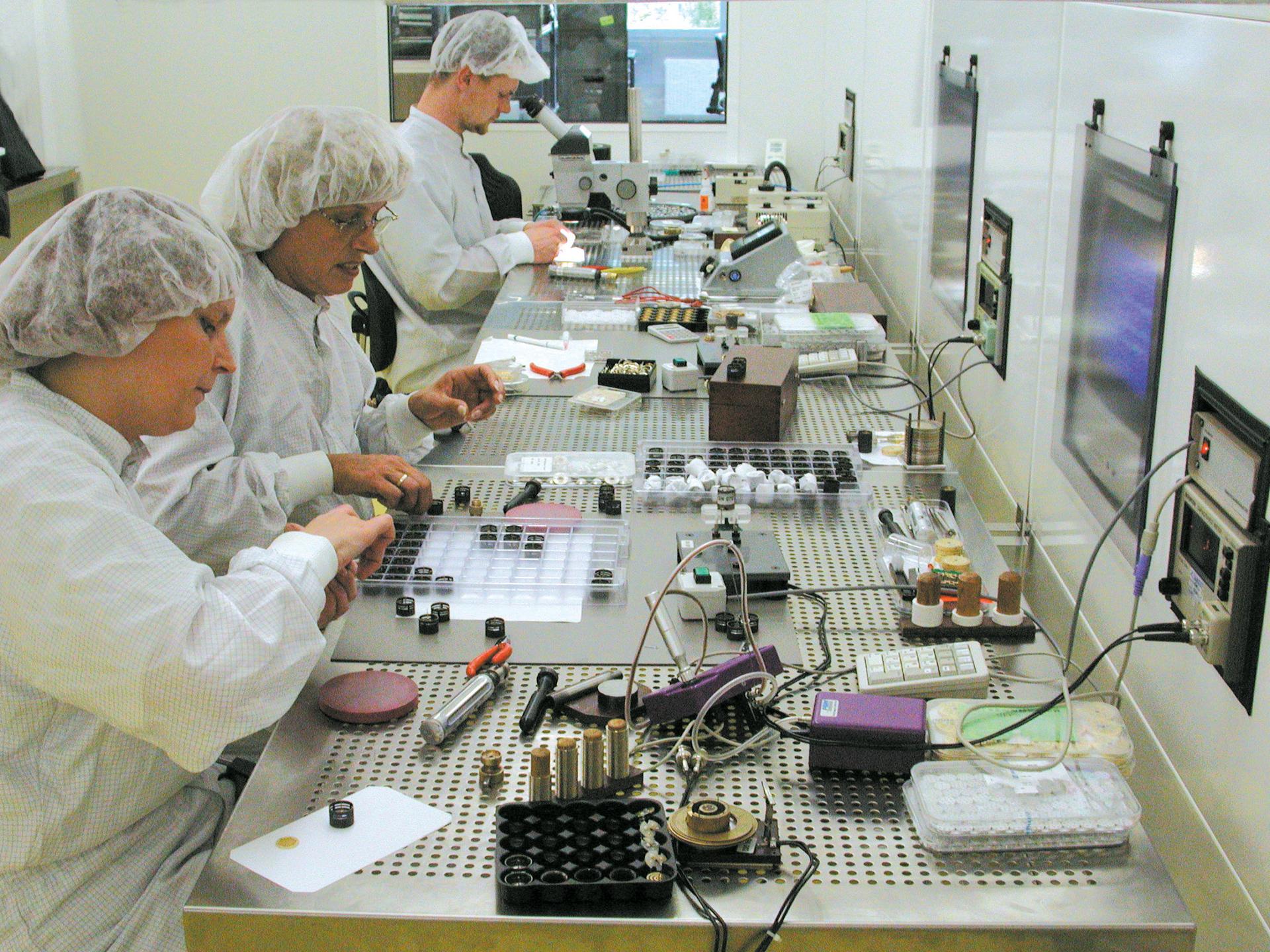 Technicians assembling electronic components at a workbench in a cleanroom environment.