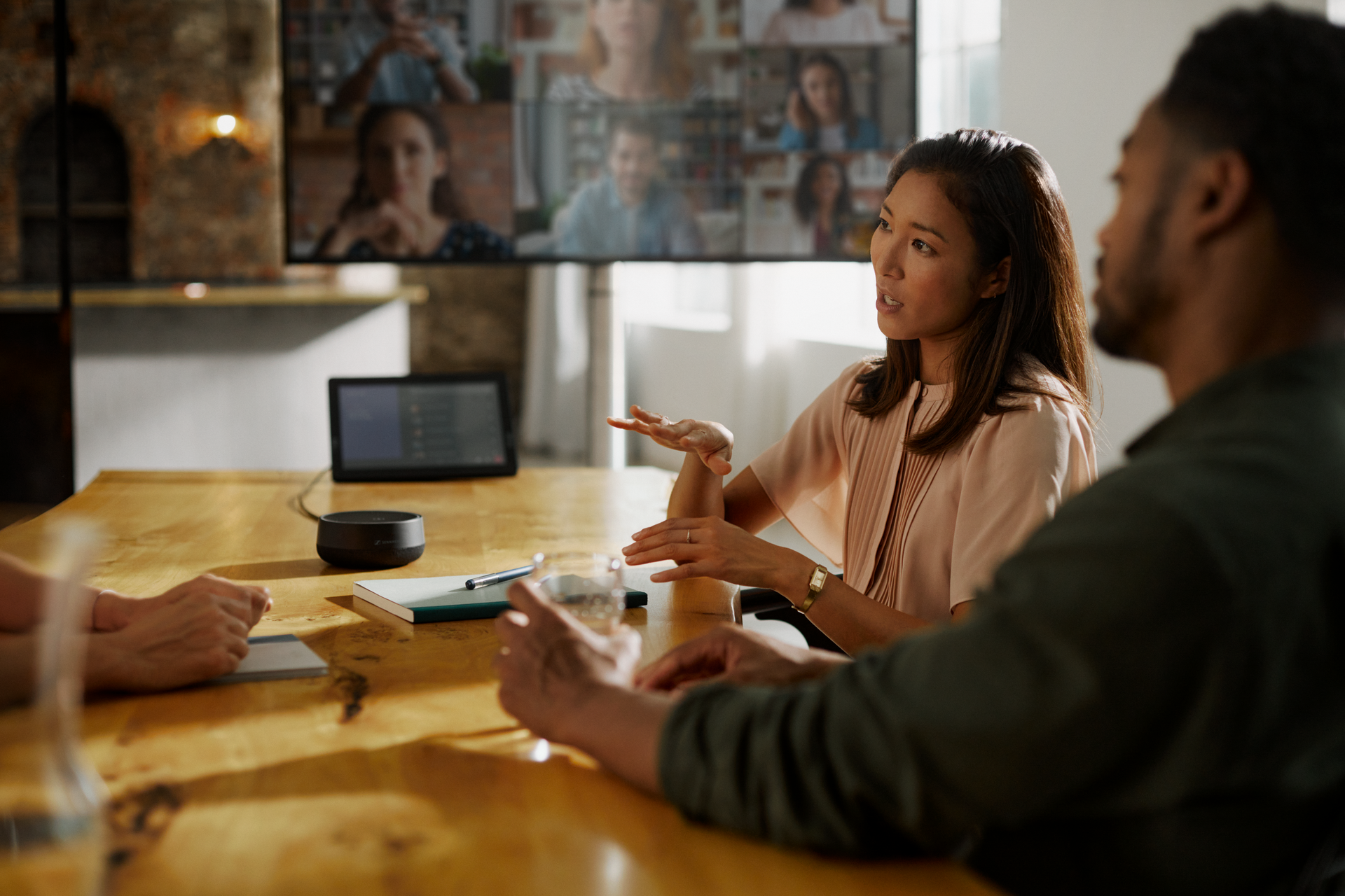 Woman leading a hybrid team meeting with a speakerphone, notebook, and screen displaying remote participants.