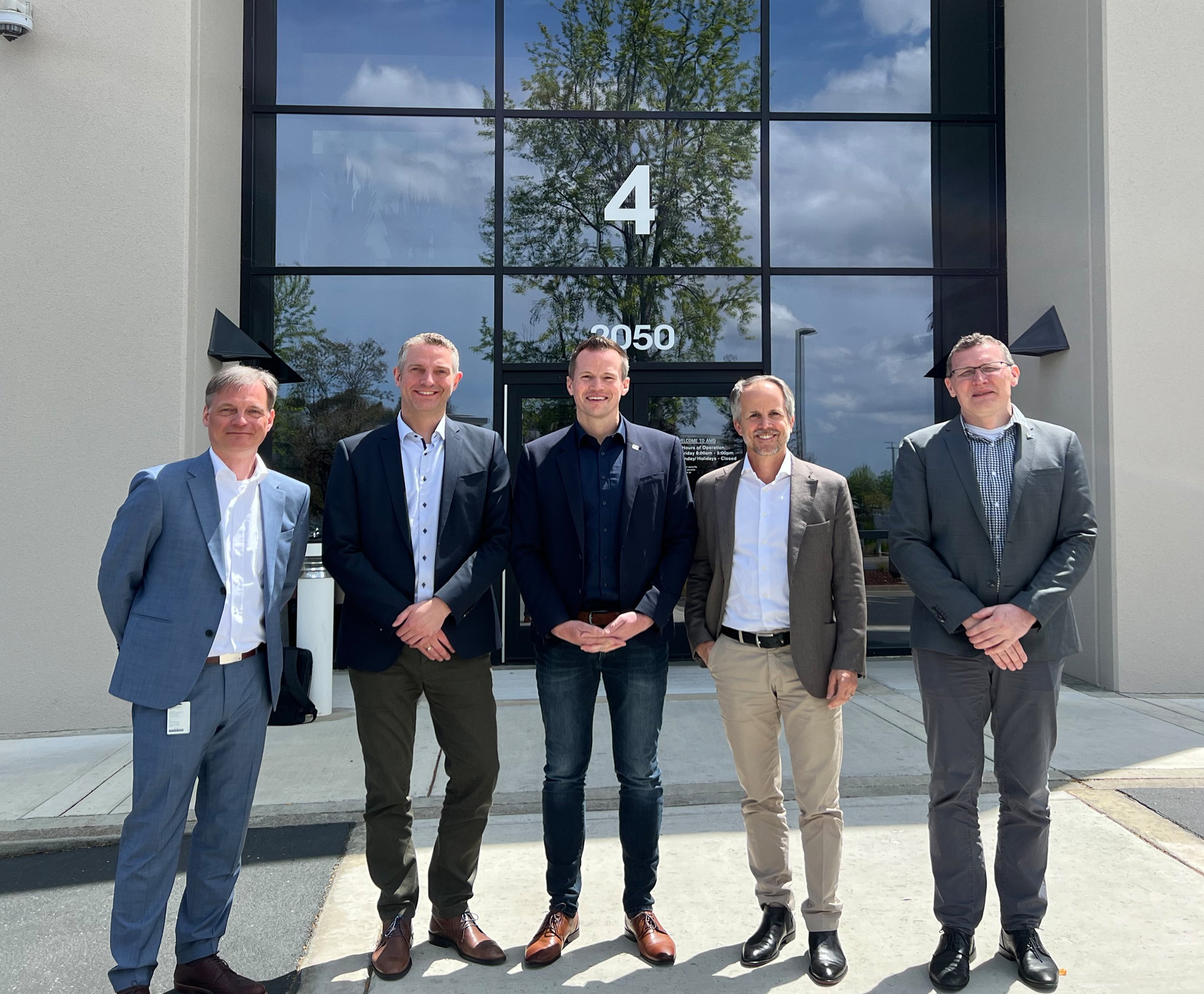 Group of five men in business attire standing in front of a building with glass doors.