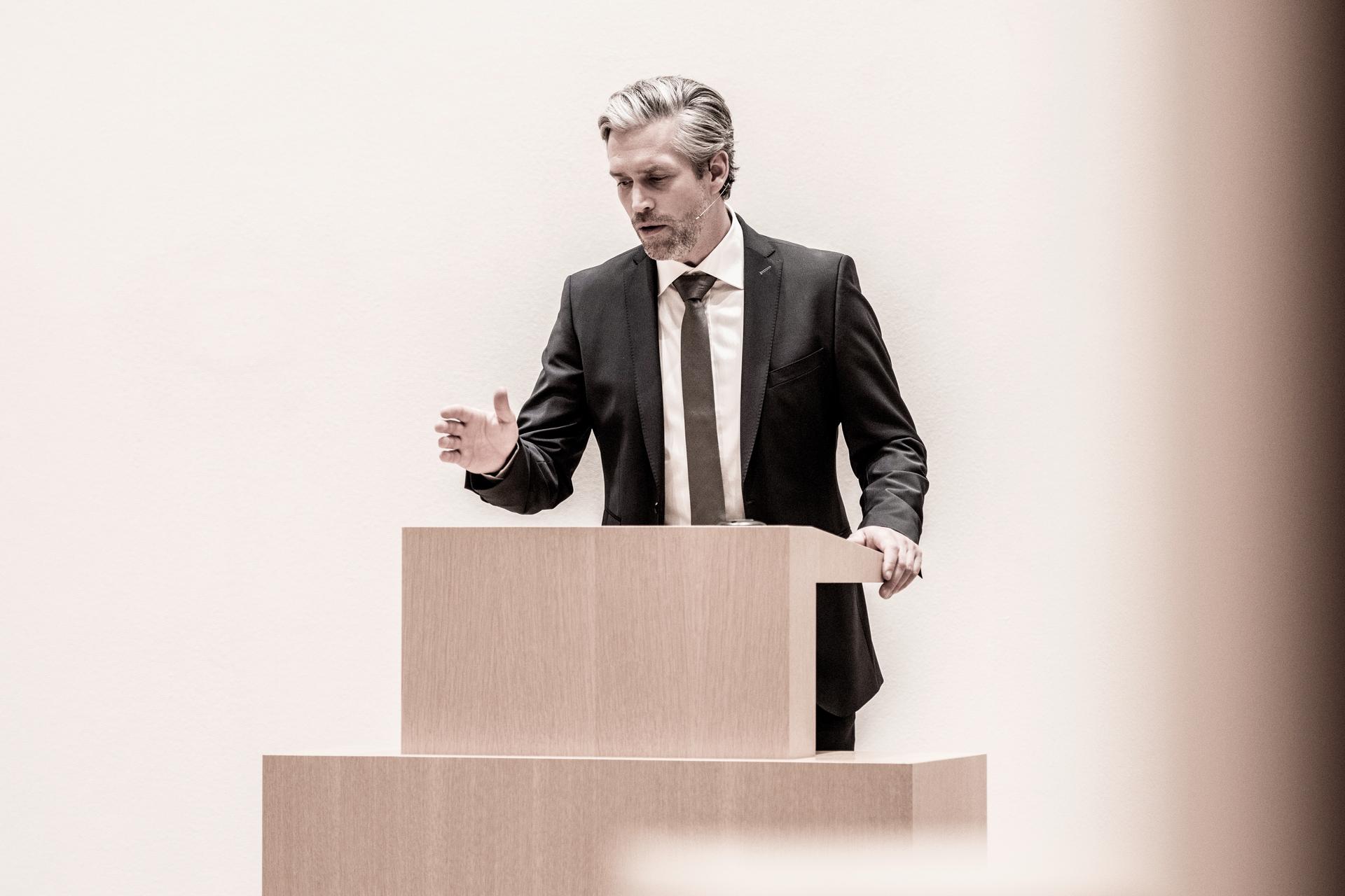 Man in a suit speaking at a wooden podium.