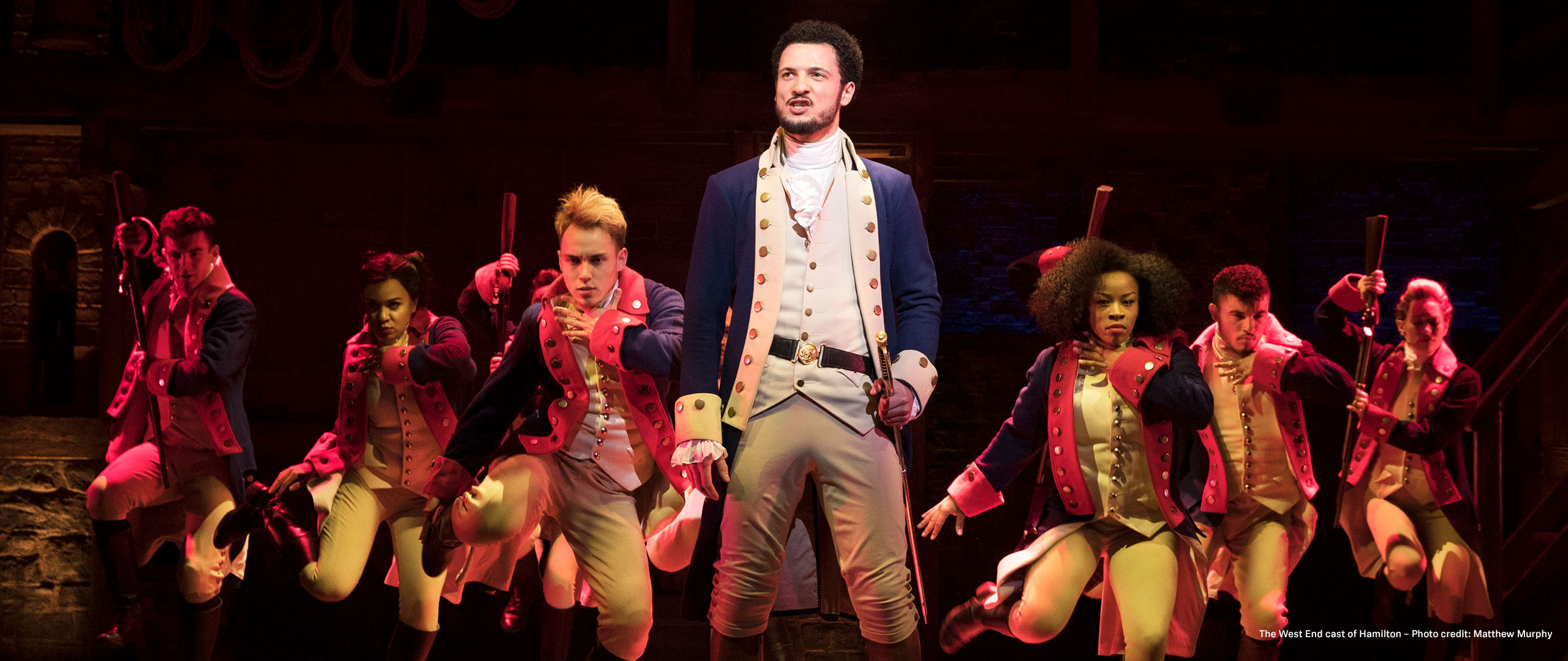 Cast of Hamilton performing on stage in period costumes.